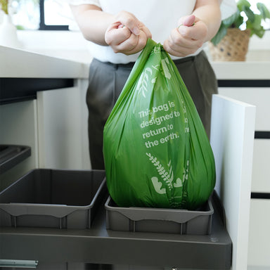 Hands tying green 27L with tie handles inside a bin in a kitchen